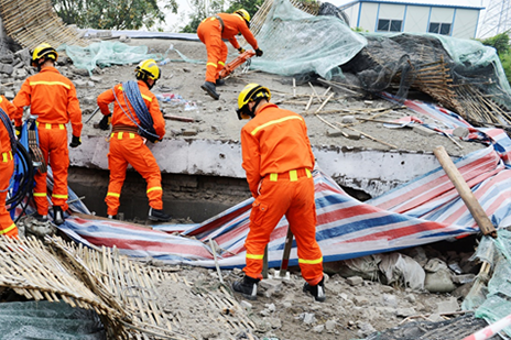 Four emergency responders in orange jumpsuits and yellow hardhats dig through rubble after a disaster